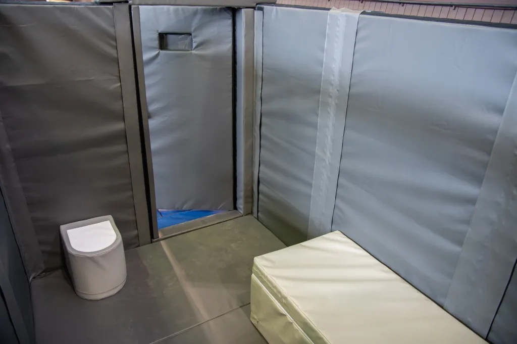 Padded custody cell interior for training with foam-wrapped PVC walls, a padded bench, and a small padded toilet.
