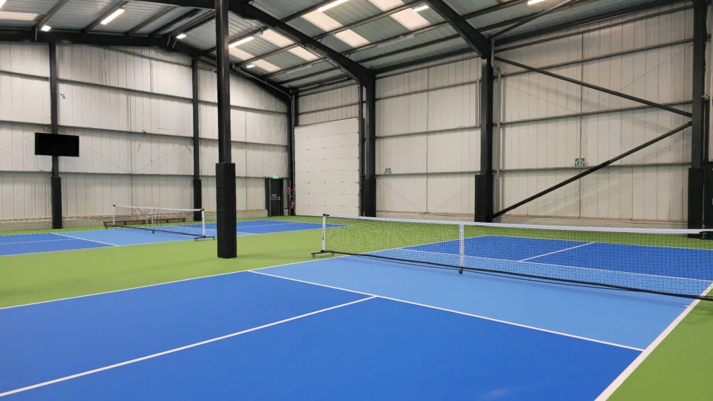 Indoor blue and green courts with black square post pads on steel posts and white nets inside a metal-roofed sports hall.