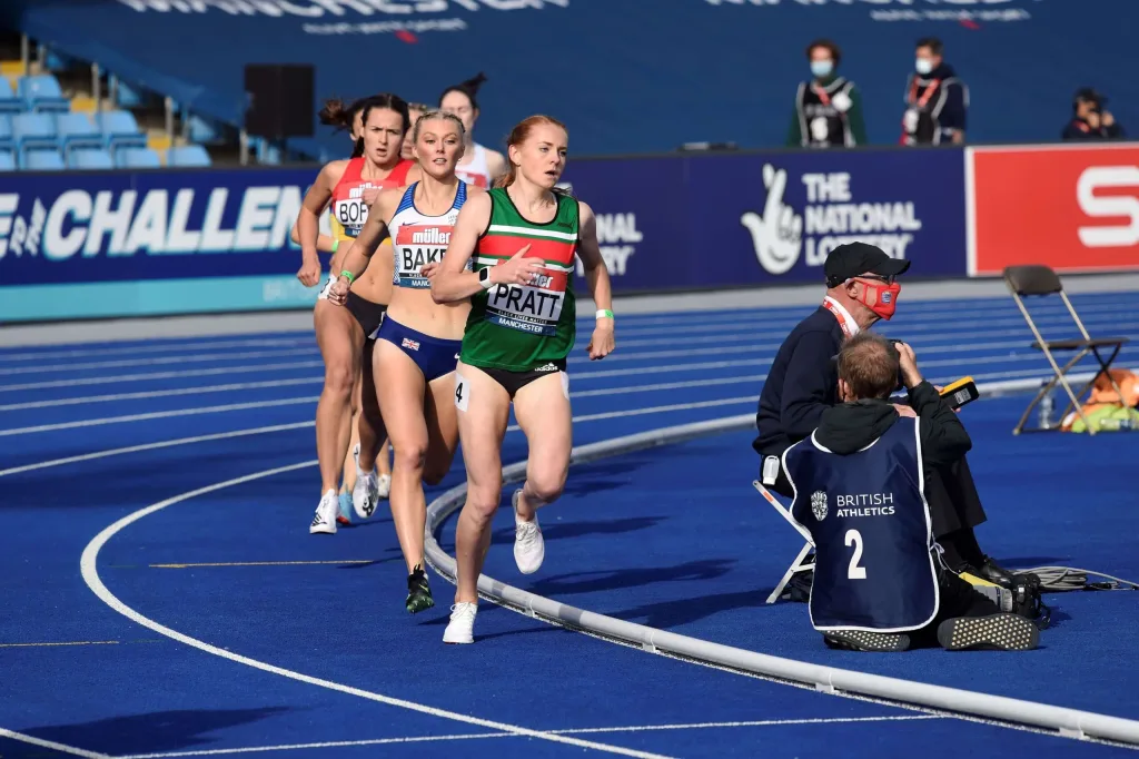 Track kerbing curving along a blue athletics track with female runners racing beside it, lead runner wearing bib 'PRATT' and officials seated nearby.