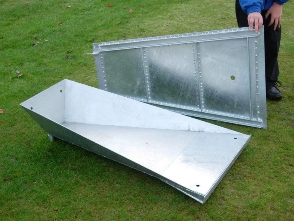 Two metal pole vault trough sections on grass, one open and one with a recessed lid leaning upright beside a person's hands.