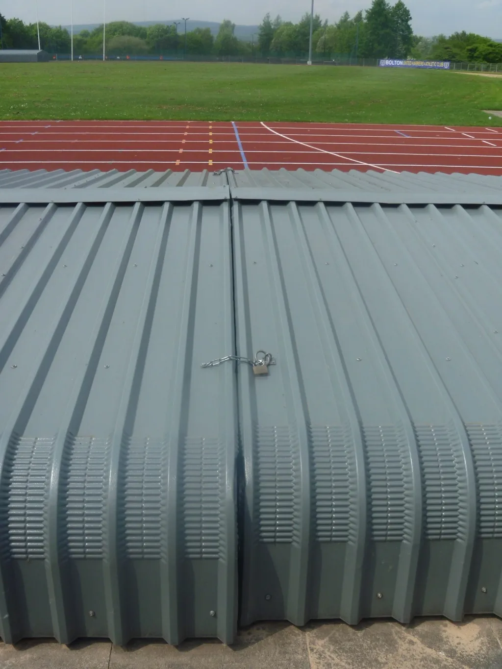 Steel wheelaway sandpit cover in two bolted sections with central padlock and chain, corrugated panels before a running track.