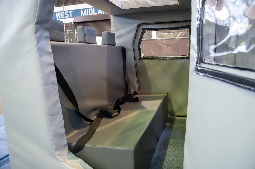 Padded training car interior with heavy-duty PVC cover, built-in bench seats and black seatbelts, small window, and background sign "WEST MIDLAND".