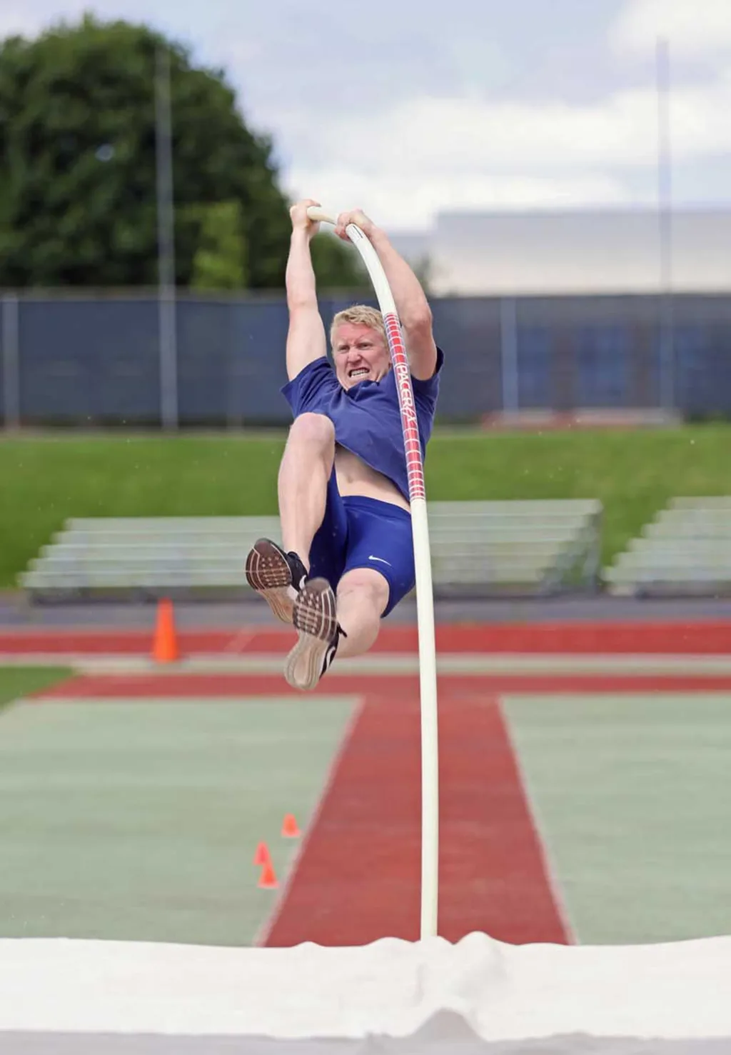 Athlete in blue shirt and shorts gripping a curved white pole while airborne above a pole vault landing mat.