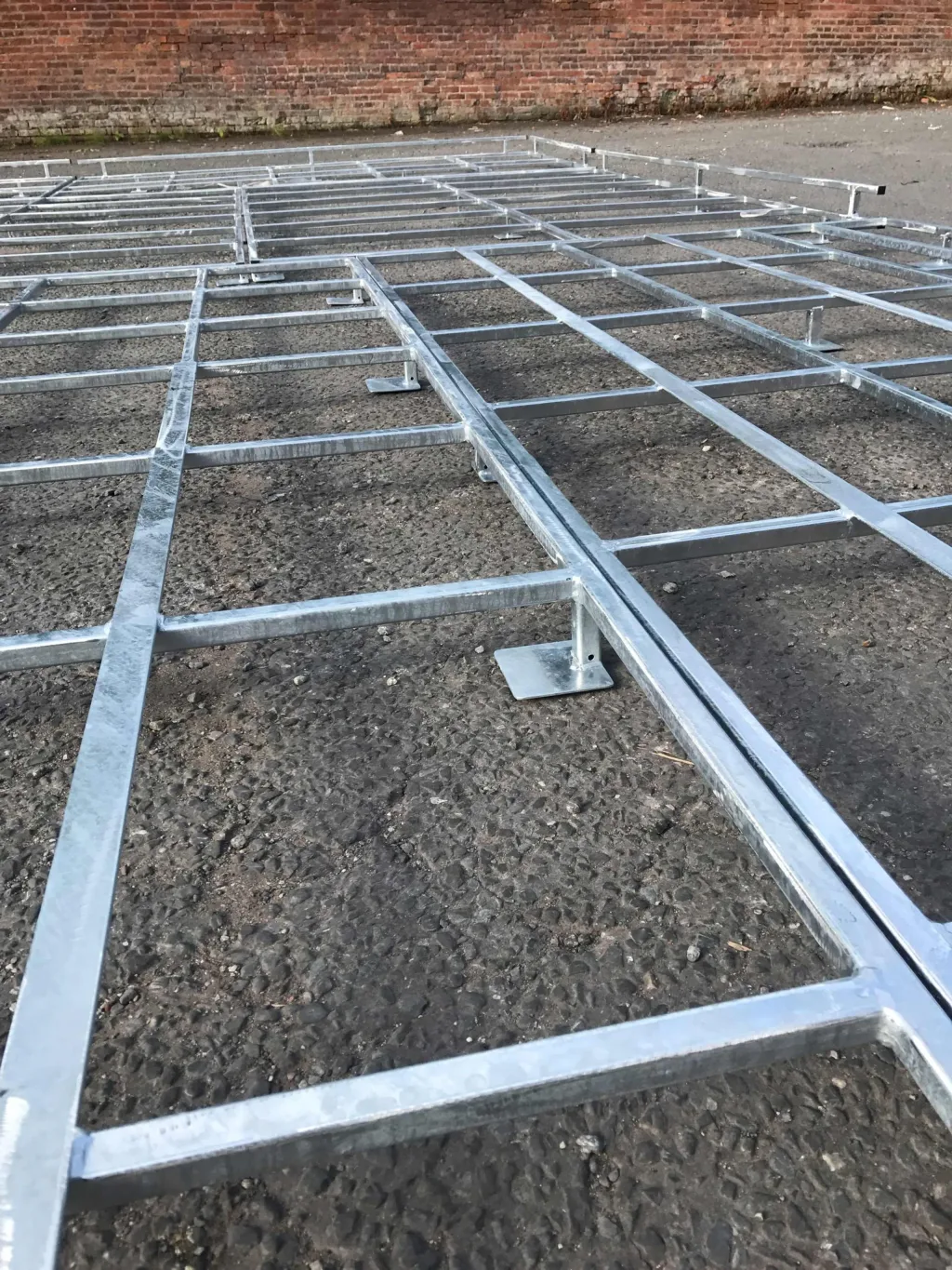 Galvanised steel platform grid with wide feet sitting on asphalt in front of a brick wall.