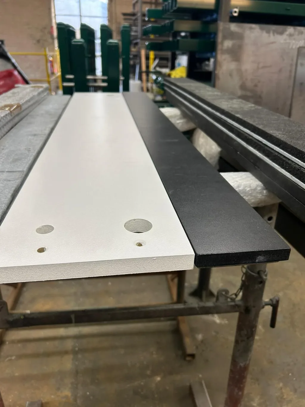 White wooden take-off board with pre-drilled holes on a galvanised steel frame next to a black wooden strip in a workshop.
