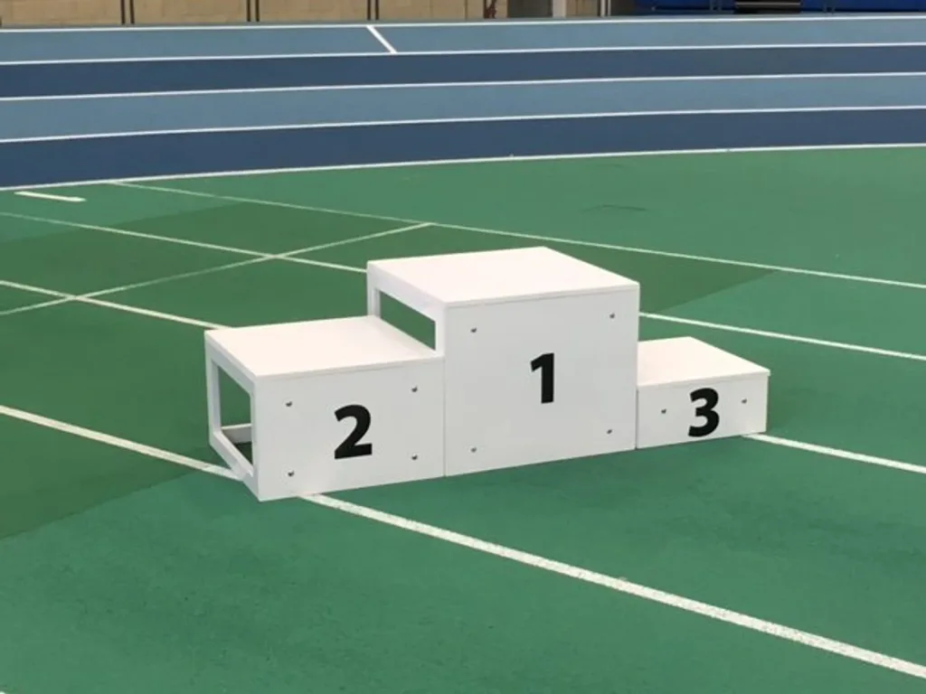White three-step winners podium labeled 2, 1, 3 placed on a green indoor athletics track with blue running lanes in the background.