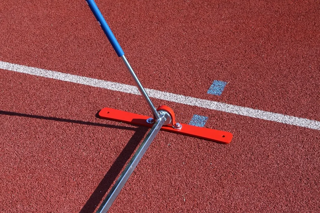Spring-back hurdle with a blue padded top bar on a galvanised steel and aluminium telescopic frame and red base plate, resting on a red track beside a white lane line.