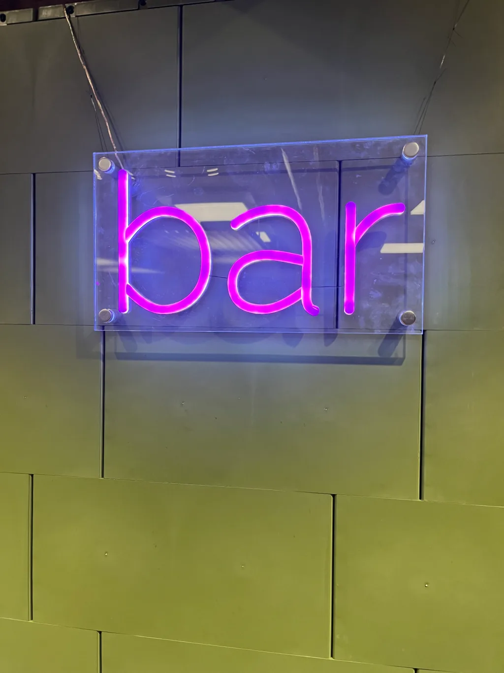 Purple neon sign reading bar mounted on a clear panel against an olive-green tiled wall.