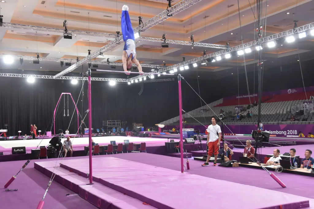 Gymnast in blue and white leotard performing a handstand on a high bar above pink landing mats, background signage reading "Palembang 2018".