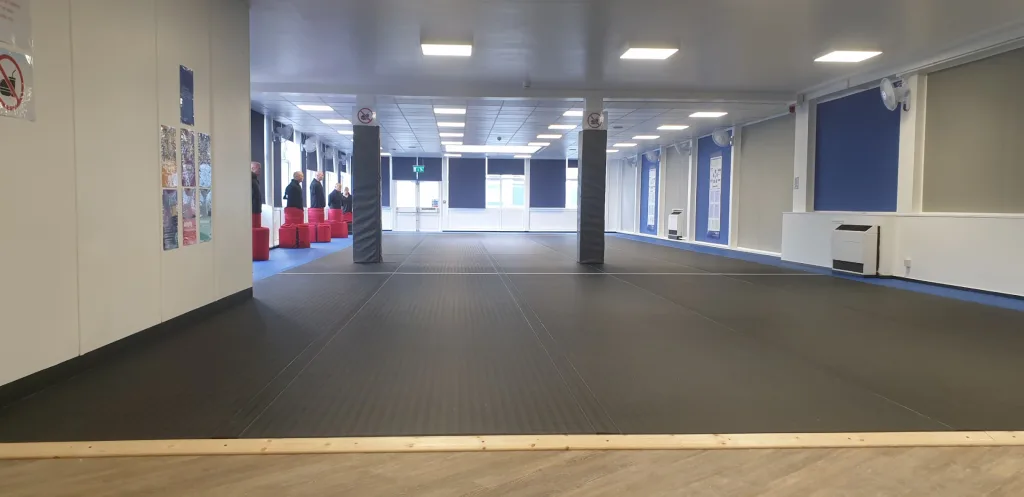 Black roll-out mats covering a large multipurpose room with blue trim, padded grey columns and red seating at the far left.