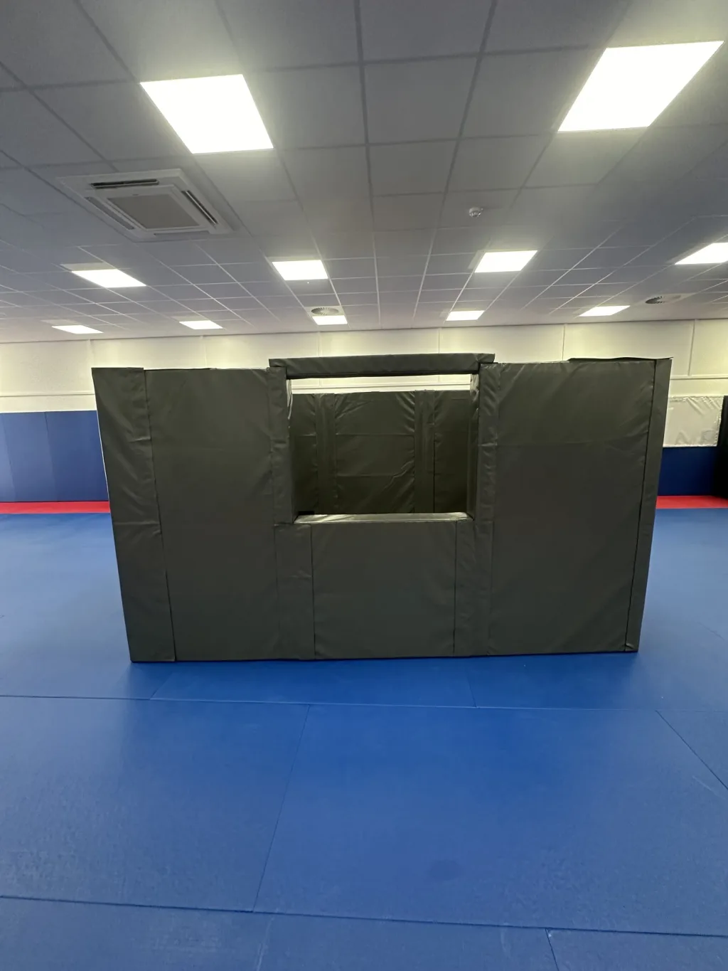 Foam and PVC custody cell with dark grey padded walls and a central window opening, set on blue mats inside a training hall.