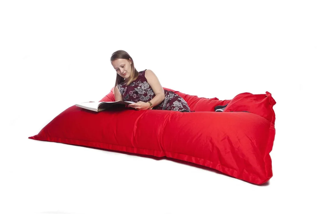 Young woman reclining on a large red cotton floor cushion, propped on her elbows and reading a book.