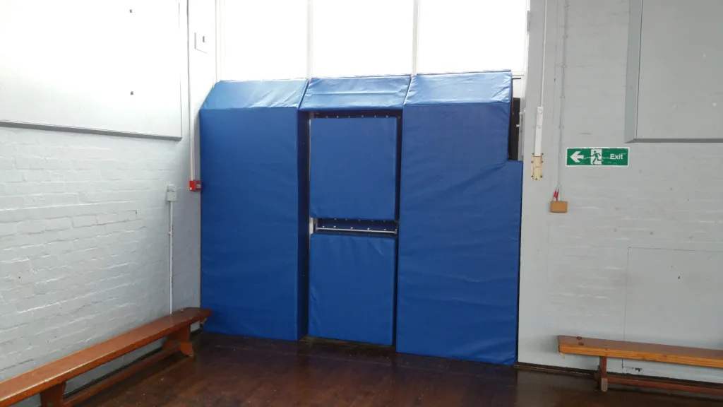 Blue wall padding panels covering a recessed doorway in a gym hall, wooden benches at either side and an "Exit" sign on the right.