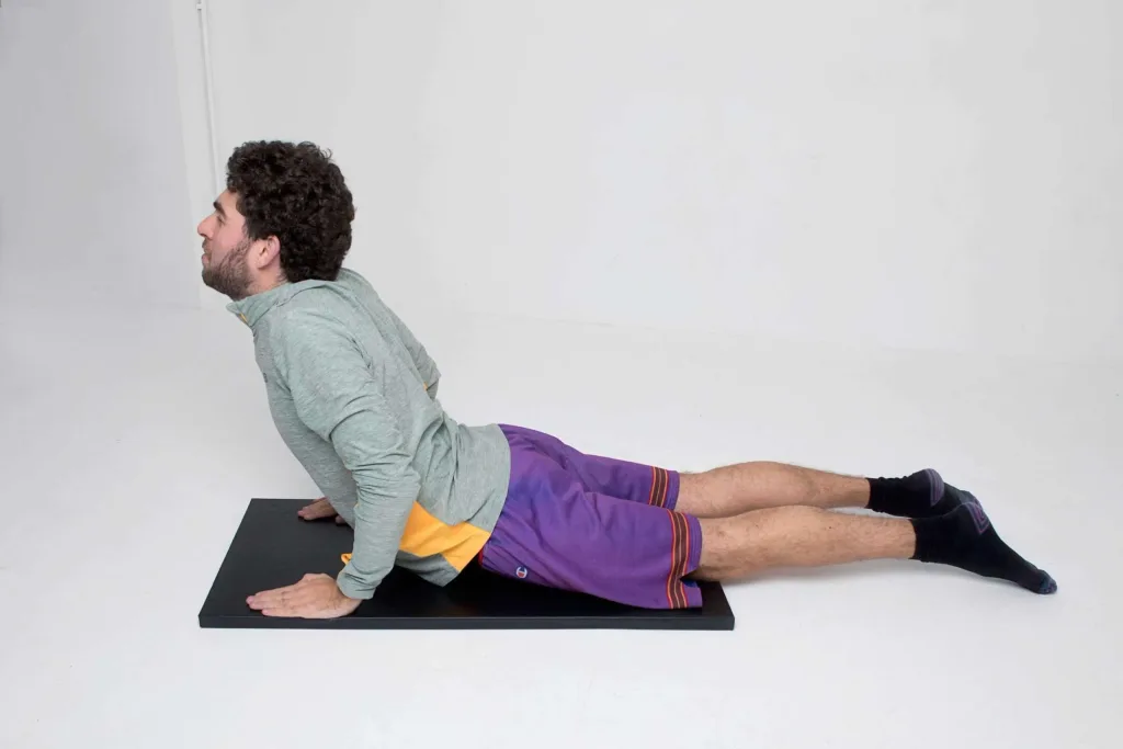 Person performing a cobra stretch on a black exercise mat in a white studio.