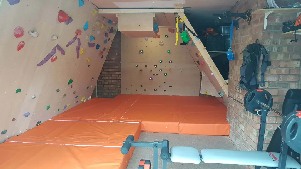 Orange PVC crash mats cover the floor under a wooden indoor bouldering wall with multicolored holds in a small brick-walled garage gym.