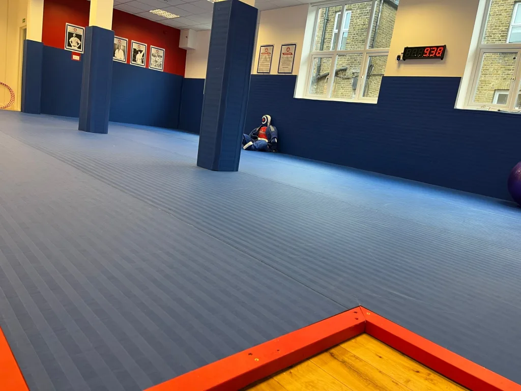 Blue PVC roll-out mat covering a martial arts studio floor with padded blue wall panels and pillars, digital clock reading 9:38, and red corner trim.