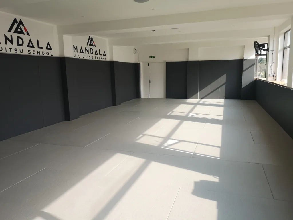 Bright training room with light grey polyester PVC combat mats and dark grey wall pads, text "MANDALA JIU JITSU SCHOOL" on the wall.