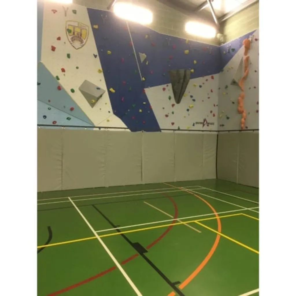 Beige hanging mats folded upright along the base of a blue-and-white climbing wall inside a gym with green multi-sport flooring.