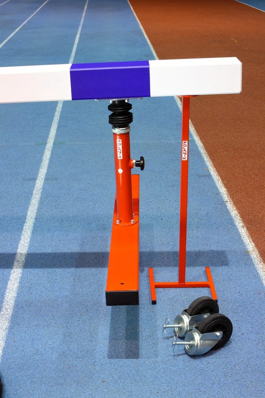 White-and-purple steeplechase hurdle mounted on orange telescopic legs with spare caster wheels on a blue indoor running track beside a brown lane.