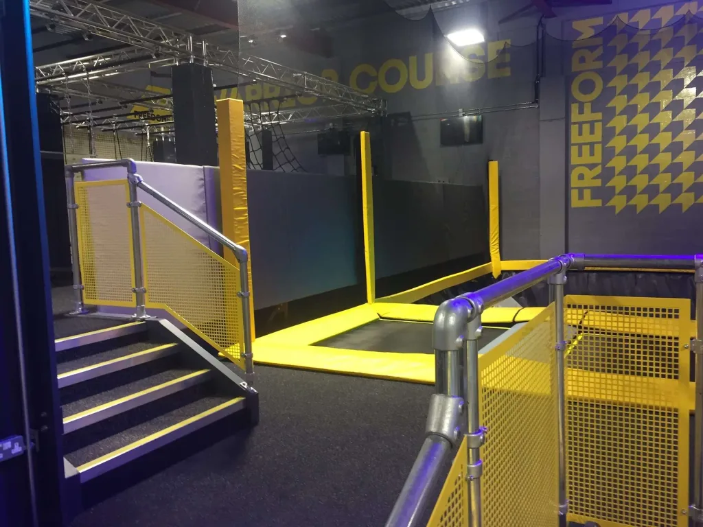 Indoor trampoline with bright yellow PVC padding, metal railings and steps in a play area, wall text FREEFORM.