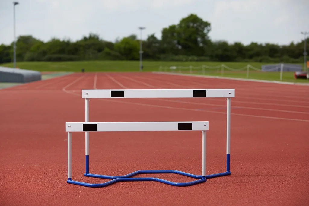 Two white track hurdles with blue curved bases positioned on a red running track with green field and trees in the background.