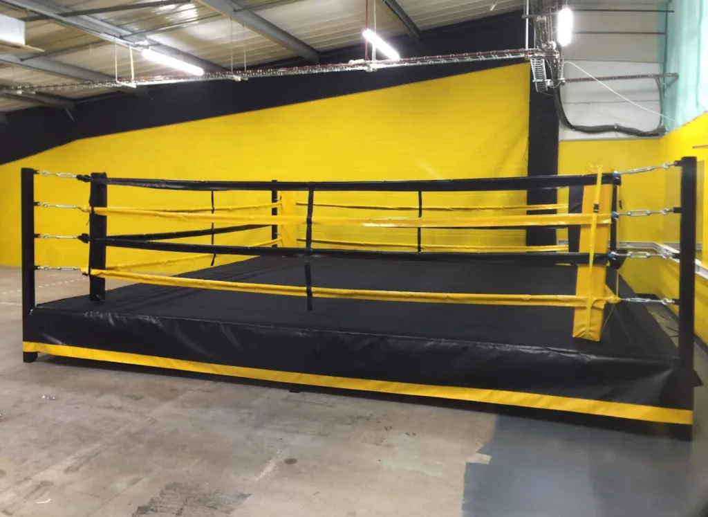 Platform training ring with black canvas and skirt, four yellow ropes and square black corner posts in a yellow-walled training space.