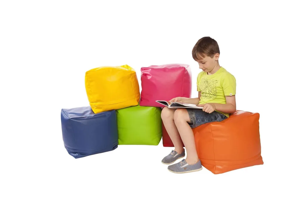 Boy sitting on an orange faux leather square bean pod reading a book beside stacked blue, green, yellow and pink faux leather cubes.