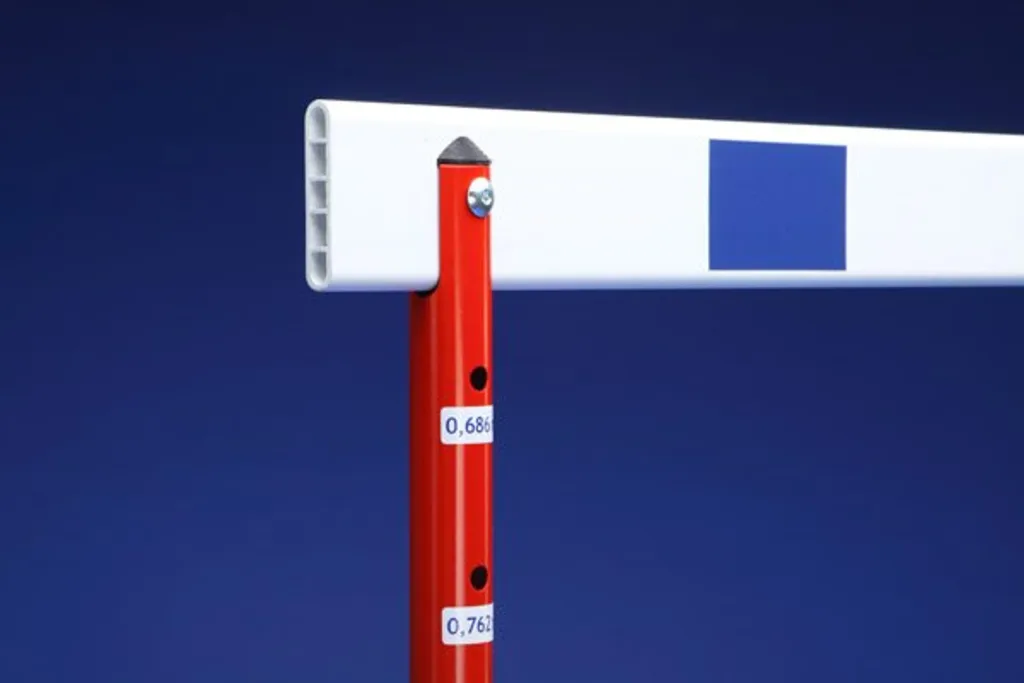 Close-up of a collapsible training hurdle showing a red steel vertical post with height labels "0,686" and "0,762" supporting a white aluminium bar marked with a blue square.