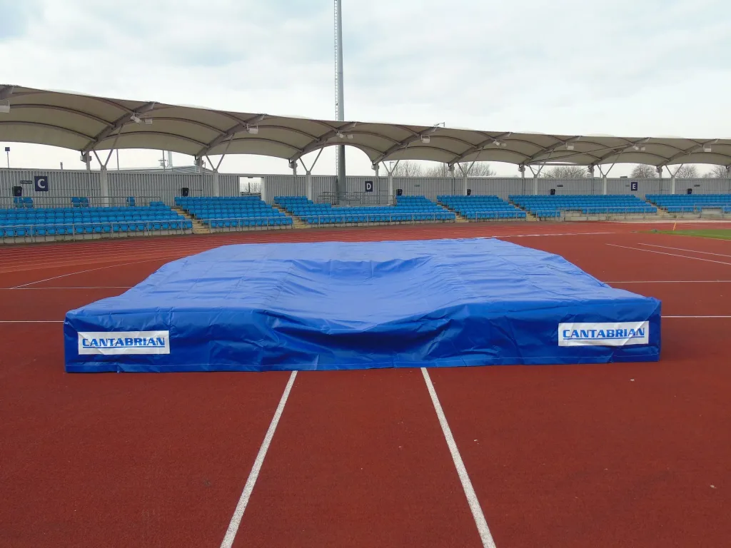 Blue PVC raincover labeled CANTABRIAN stretched over a pole-vault landing mat on a red track with empty blue stadium seats behind.