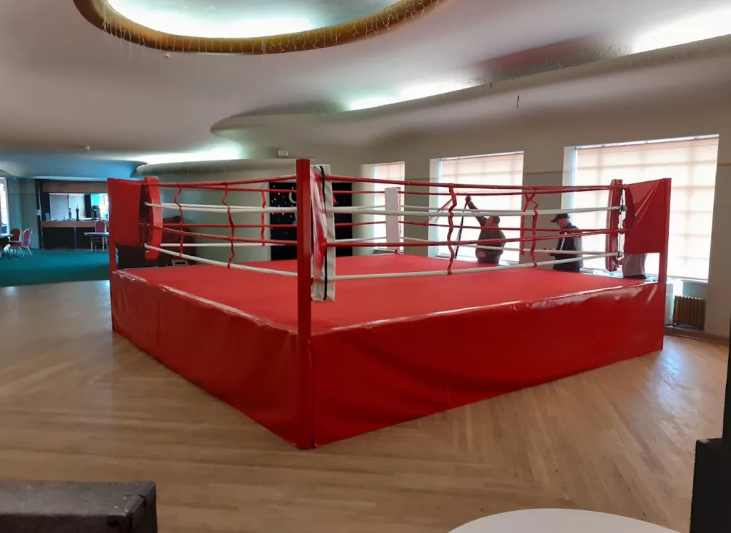 Red competition ring with white ropes and padded red corners on a raised deck placed on a wooden floor in a room with windows.