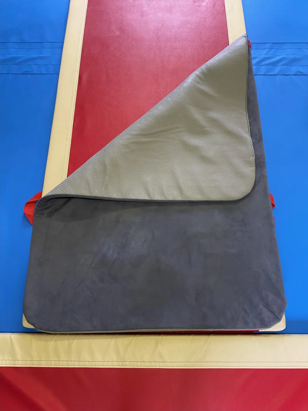 Jersey balance beam mat folded at one corner showing grey anti-slip underside and PU foam core, red top with beige trim on a blue mat beneath.