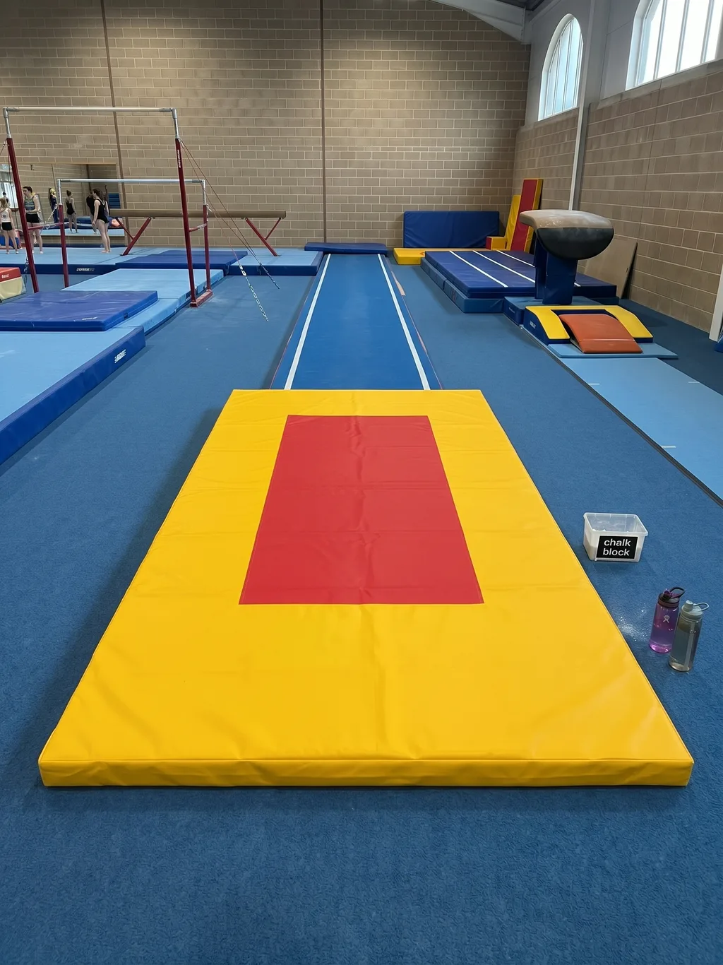 Bright yellow gymnastics mat with red target in the center at the end of a blue runway in a gym, small box labeled "chalk block" visible to the right.