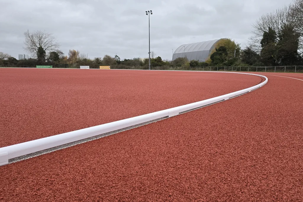 White track kerbing curving across a red athletics track surface with trees and a gray sports dome in the background.