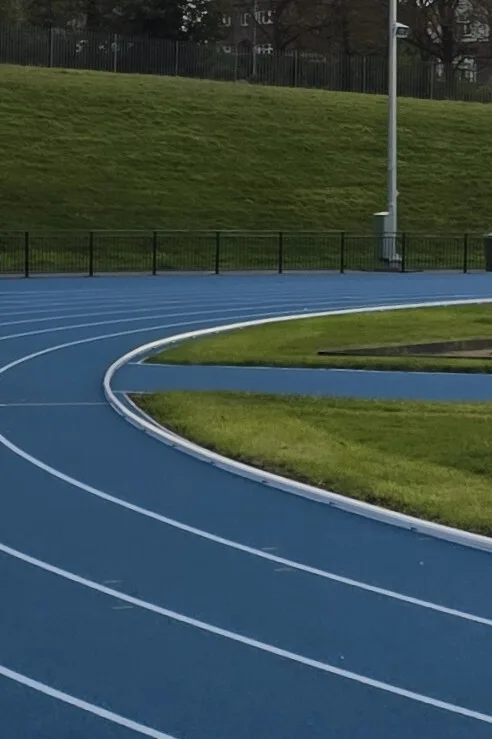 Curving blue running track with white lane lines and low kerbing bordering a grassy infield, with a fence and lamp post in the background.