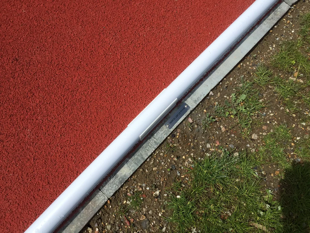 White kerb runs along a red rubber running track beside a metal edge, gravel and patchy grass.
