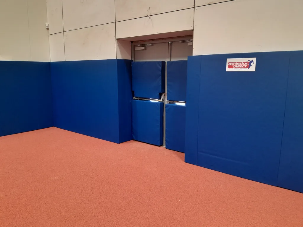 Blue polyester PVC wall pads cover a corner and double doors in an indoor gym with red rubber track flooring.