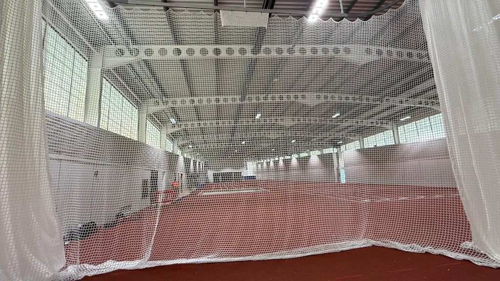 White mesh netting of an indoor throws cage in foreground, framing a red track and high industrial ceiling.