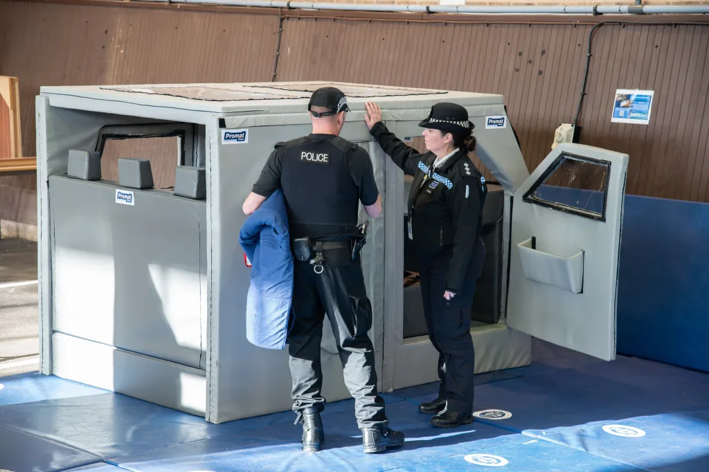 Two police officers stand beside a gray padded training mock vehicle on blue mats, one officer's vest reading POLICE.