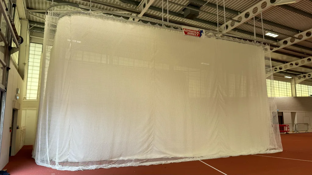 A white netted throws cage suspended from the ceiling in an indoor athletics hall, enclosing a section of red track flooring.