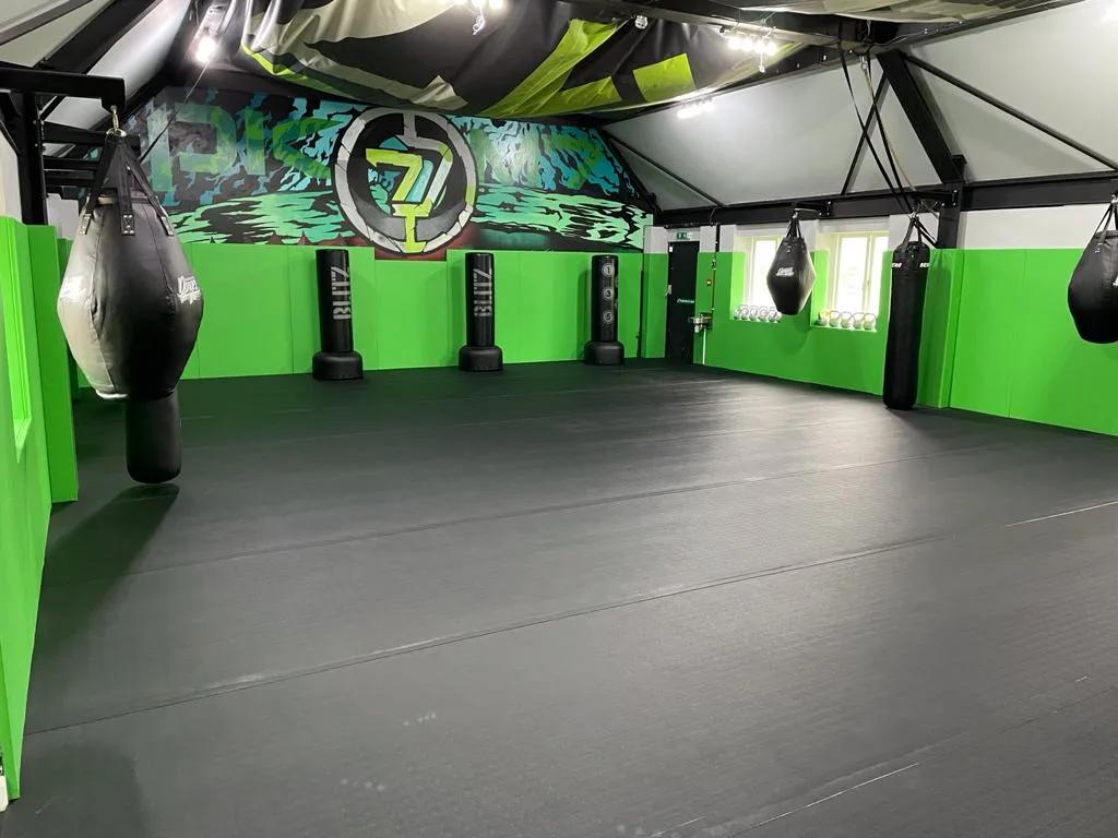 Spacious gym with black floor and bright green wall pads, hanging heavy bags, standing dummies, and mural on back wall.